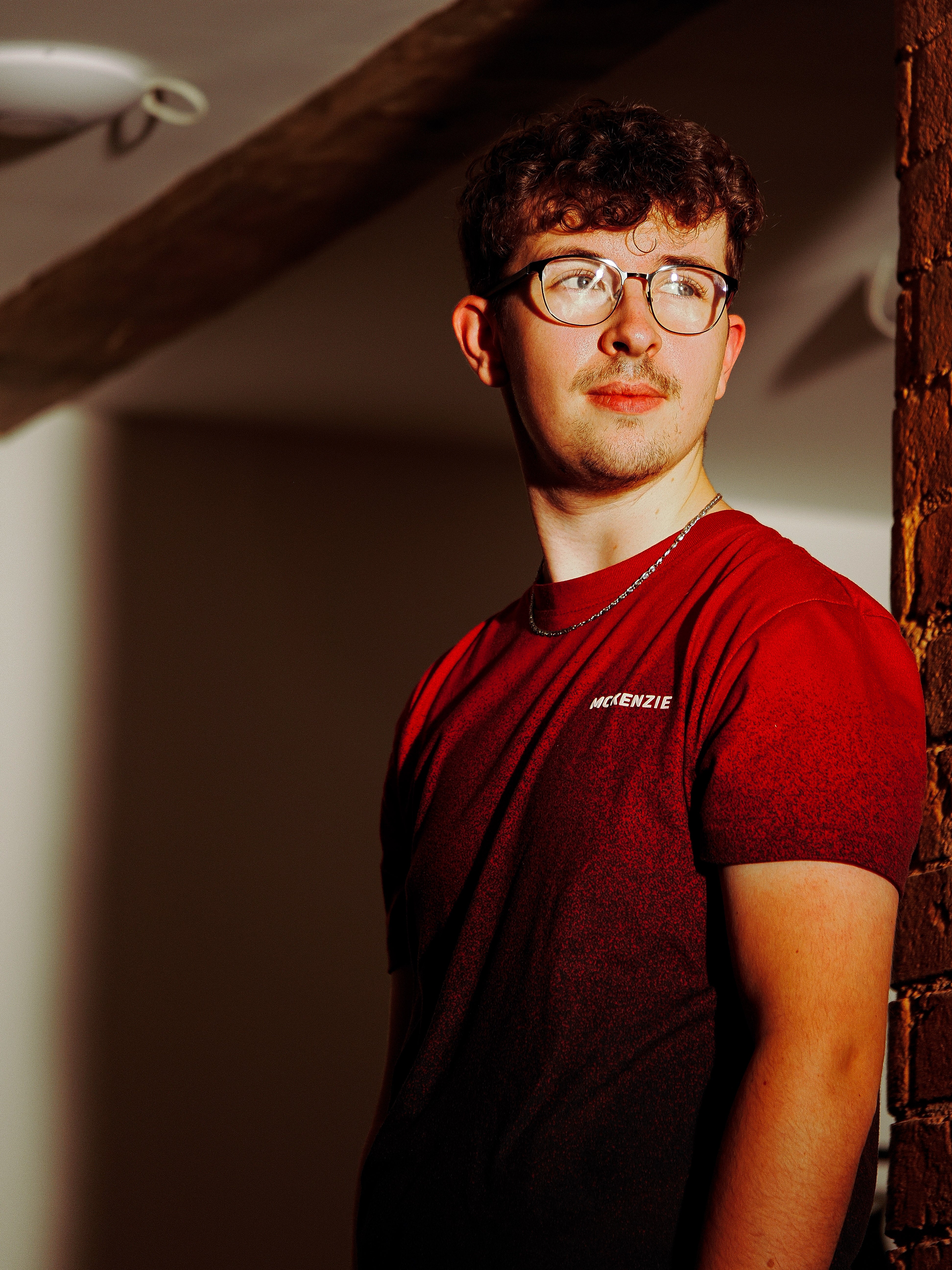 Man wearing glasses and a red shirt standing indoors with red brick walls.