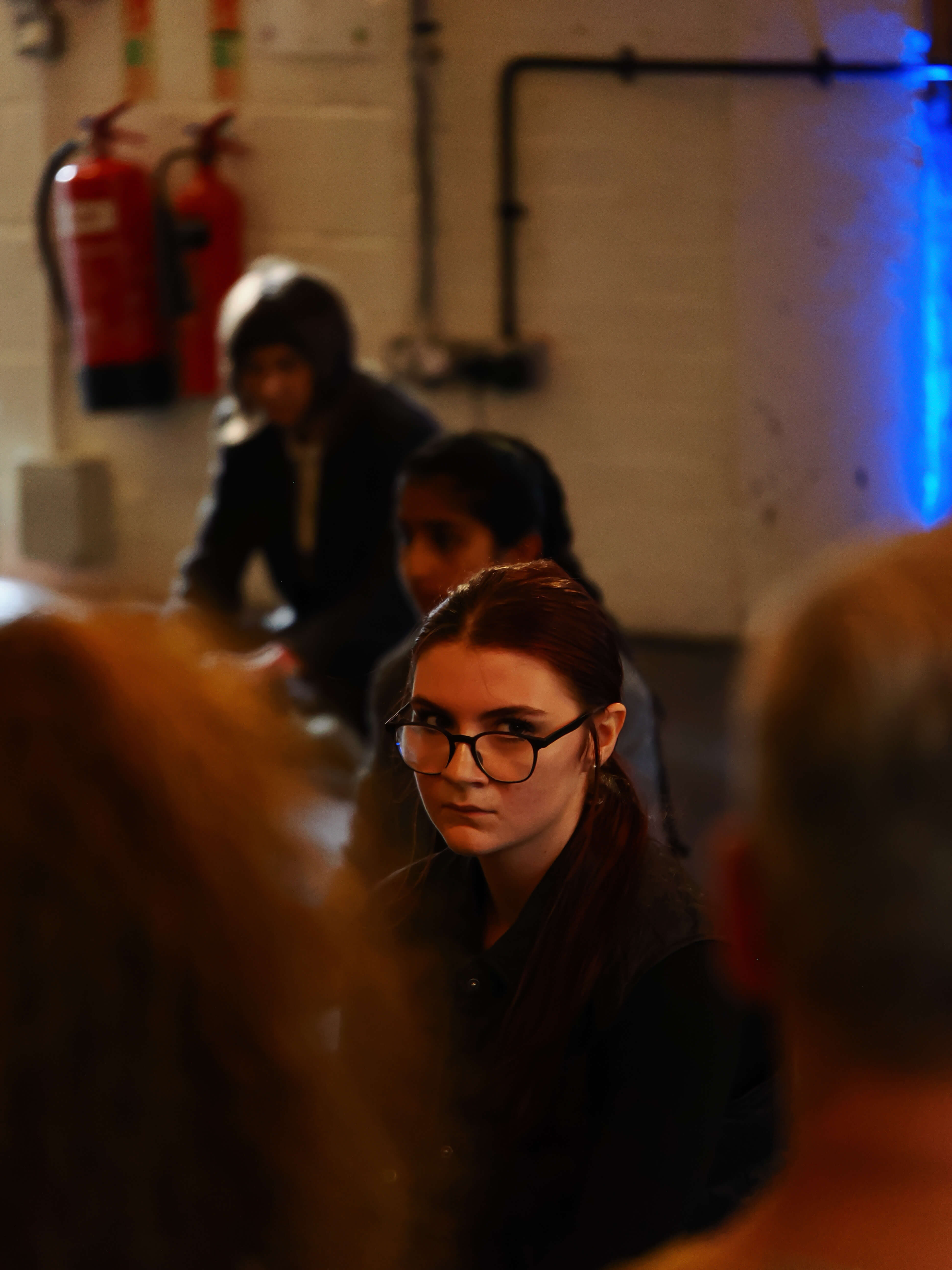 Girl wearing glasses in a room with other people and visible fire extinguishers on the wall.
