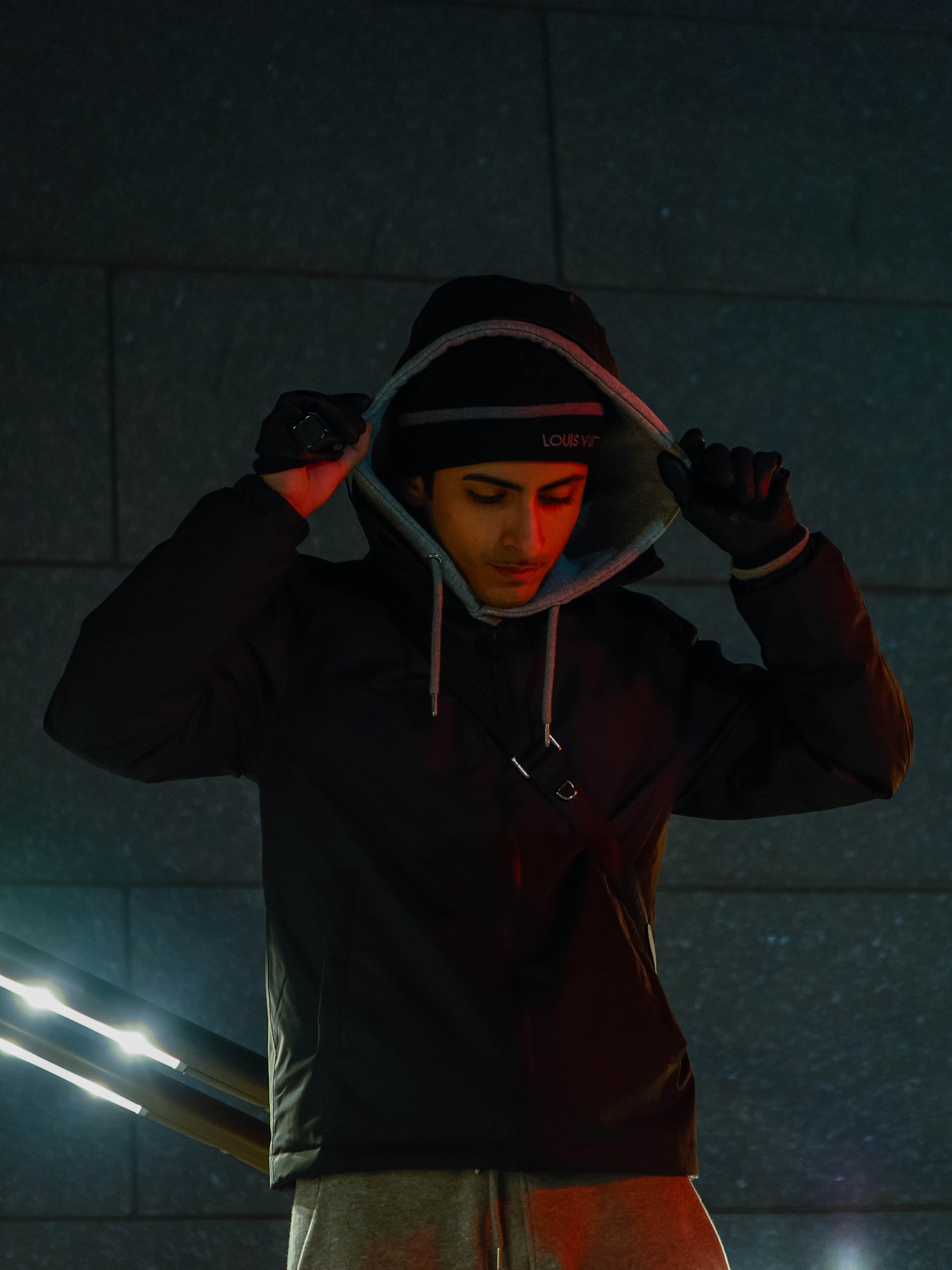 Male model wearing a dark hoodie with a hood, standing against a textured wall with a red light on his face.