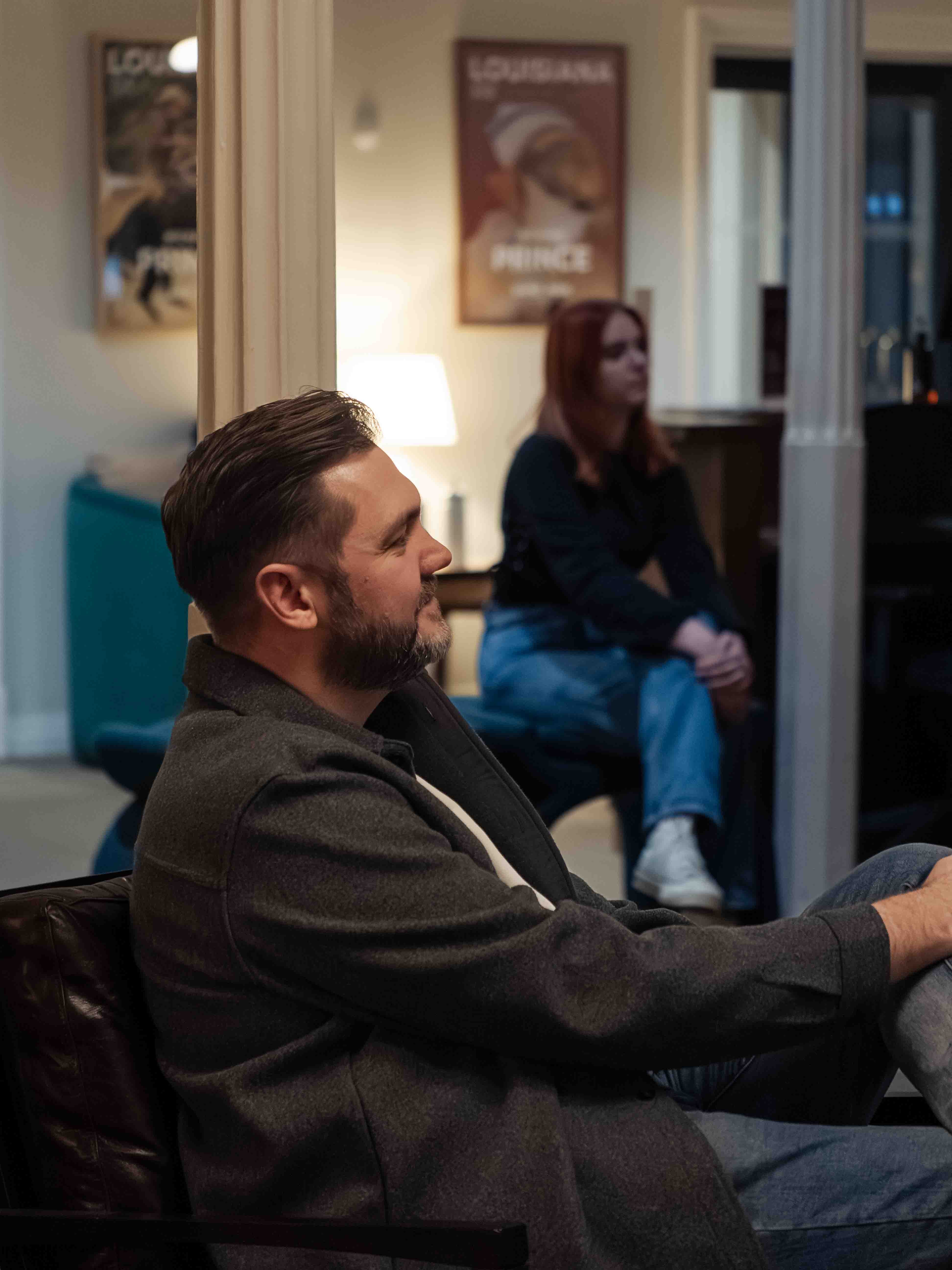 Man sitting on a couch with a woman in the background in a room with posters on the wall.