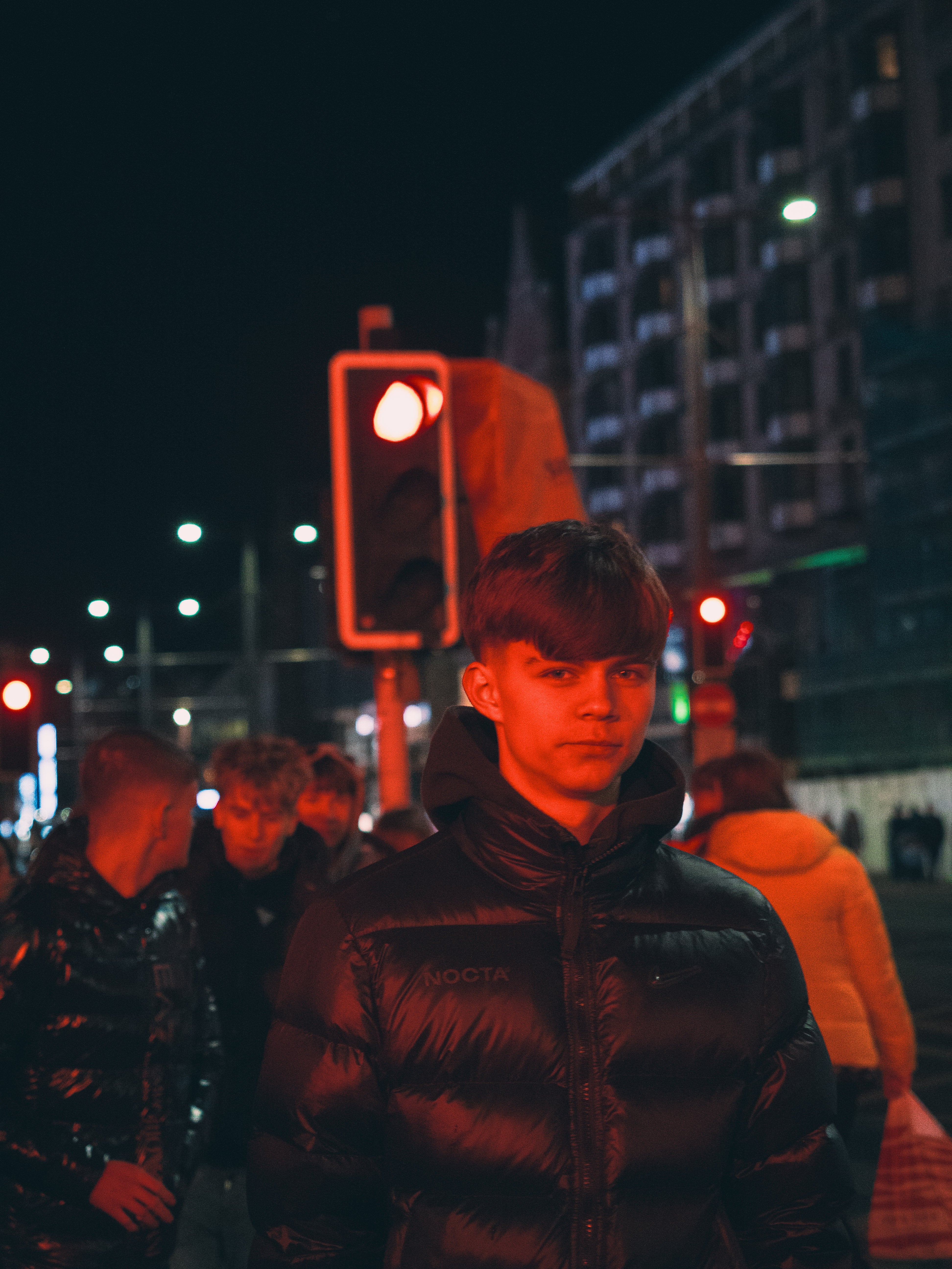 Person wearing a black jacket in an urban setting at night with red traffic lights.