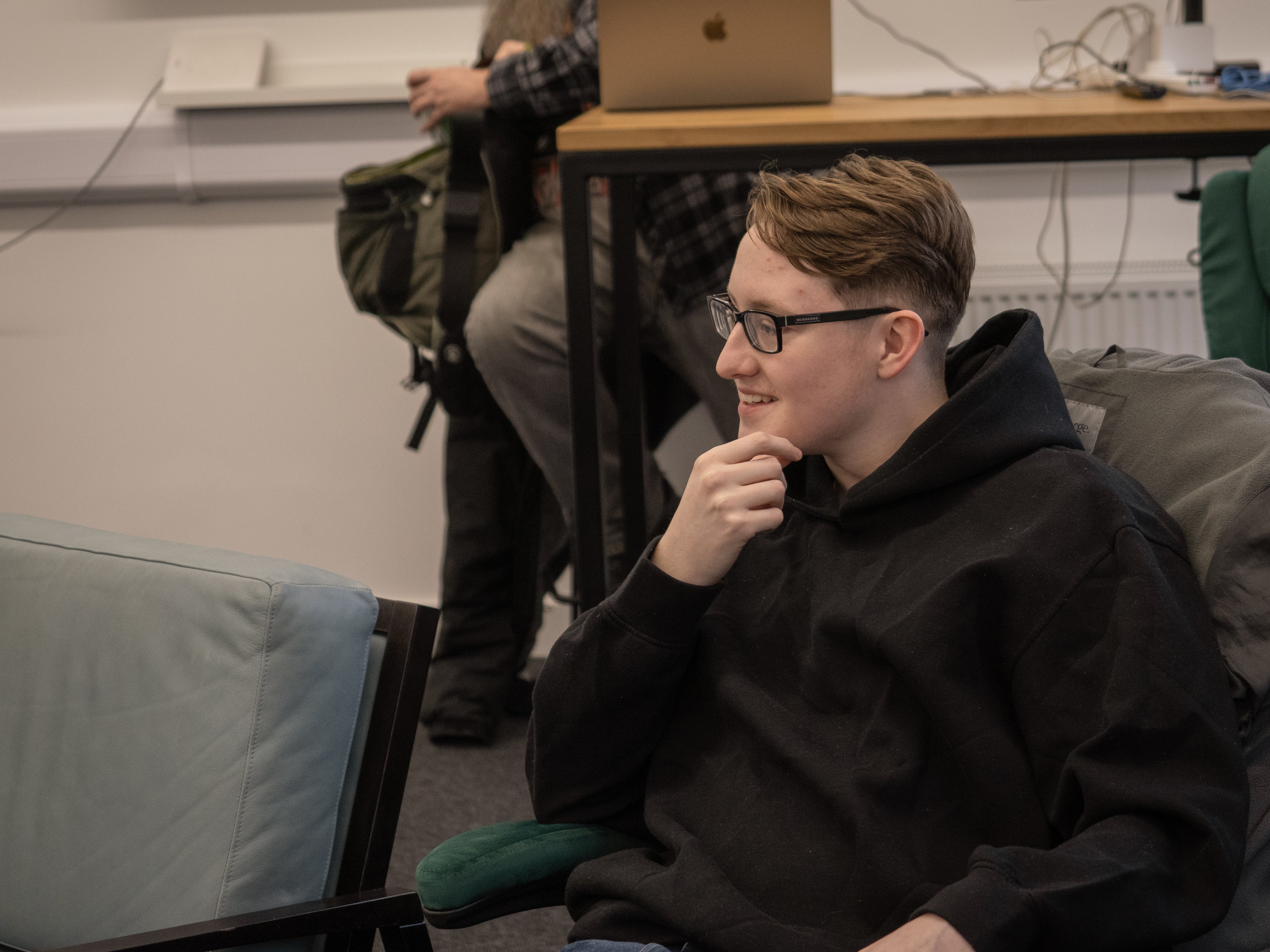 Person wearing glasses and a black hoodie sitting in a room with others and furniture.