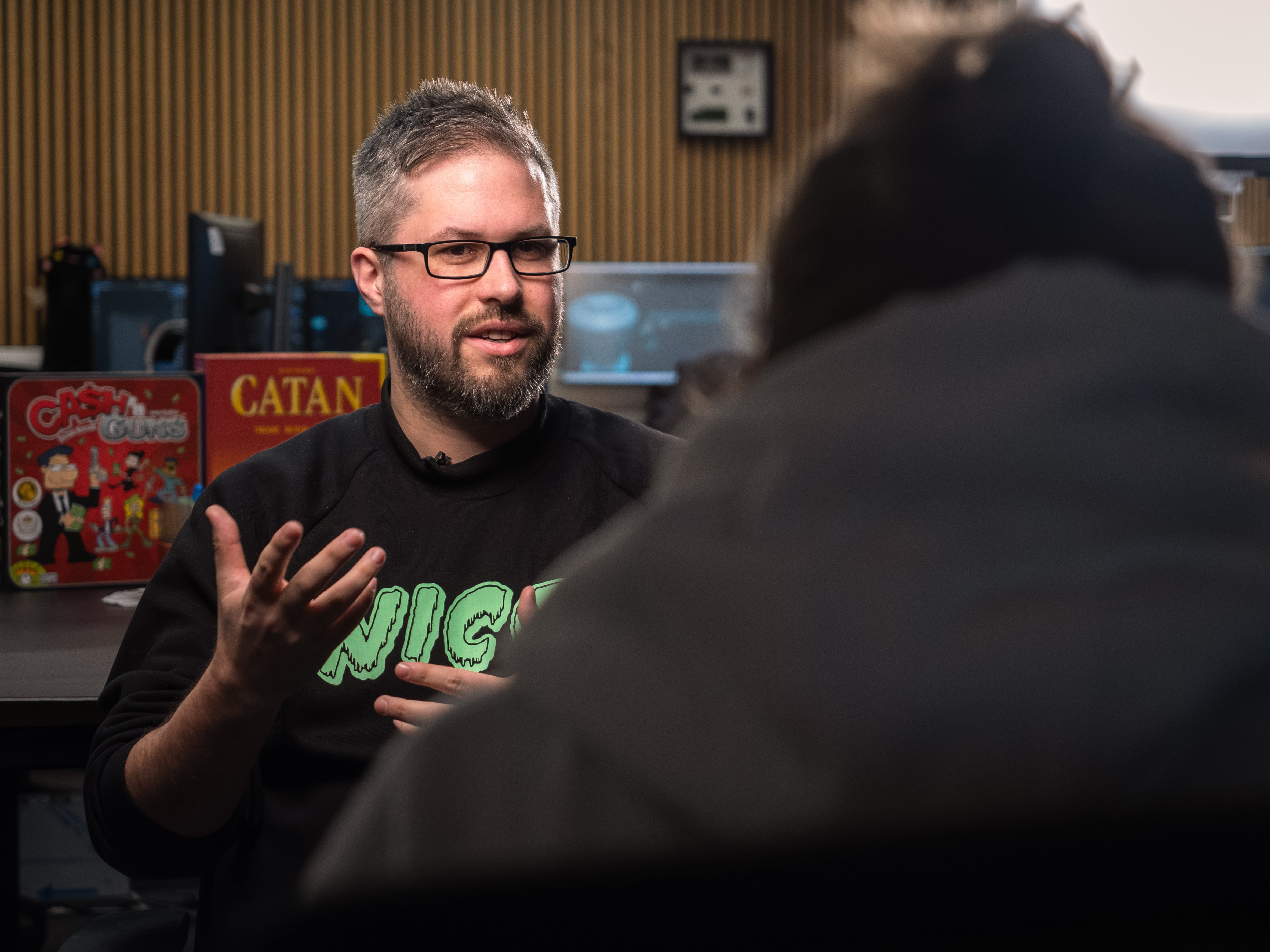 Man in a dark shirt with 'NICE' on it talking to someone in a room with gaming equipment.